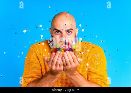 bell'uomo con raindow in barba e maglione giallo che soffia i dolciumi in baclground blu studio Foto Stock