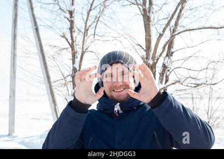 L'uomo caucasico con una barba in una giacca tiene le mani vicino al viso e ride. Passeggiata in una giornata invernale nevosa. Foto Stock