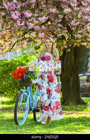 Stagione Sakura. Concetto di turismo. Trasporto e viaggi. Donna con bouquet tulipani. Sakura fiore albero. Romantico giro in bicicletta. Abito lungo ragazza retrò Foto Stock