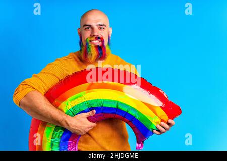 uomo con barba colorata che tiene grande palloncino arcobaleno su sfondo bludio Foto Stock