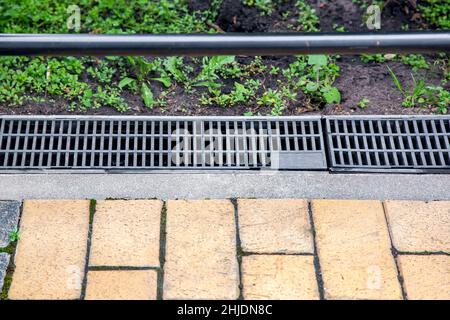 grata grigia del sistema di drenaggio per il drenaggio dell'acqua piovana nel cortile sul bordo del marciapiede da mattonelle di pietra con erbacce verdi nel terreno, paesaggistico Foto Stock