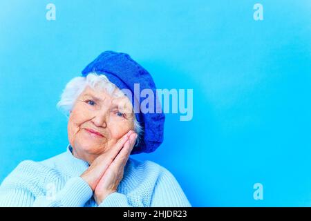 bella donna in pensione in beretto blu e caldo pullover guardando la macchina fotografica e il sorriso in sfondo blu parete in studio Foto Stock