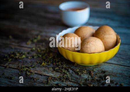 Sode uova di tè cinesi speziate in marmo su un rustico tavolo di legno con foglie di tè e una tazza di tè cinese sullo sfondo. Foto Stock