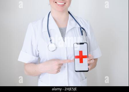 Medico femminile irriconoscibile tiene uno smartphone con una croce rossa sullo schermo. L'infermiere sorride e punta allo schermo di un telefono cellulare. Foto Stock