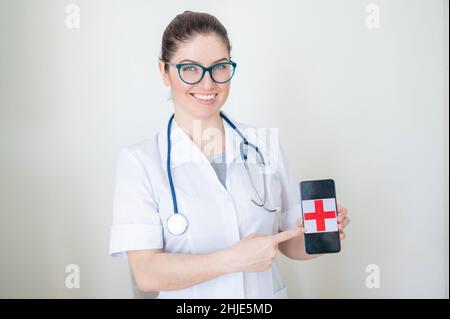 Medico femminile in occhiali tiene uno smartphone con una croce rossa sullo schermo. Un'infermiera sorride in un cappotto medico con uno stetoscopio sulle spalle Foto Stock