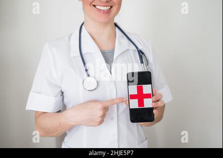 Medico femminile irriconoscibile tiene uno smartphone con una croce rossa sullo schermo. L'infermiere sorride e punta allo schermo di un telefono cellulare. Foto Stock