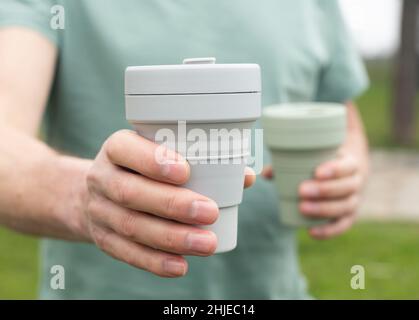 Mano maschio con due tazze eco riutilizzabili in mano, dando e offrendo tazza di caffè grigio silicone in avanti. Foto Stock