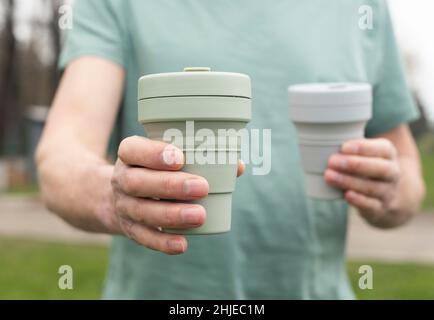 Mano maschio con due tazze eco riutilizzabili in mano, dando e offrendo tazza di caffè verde silicone in avanti. Foto Stock