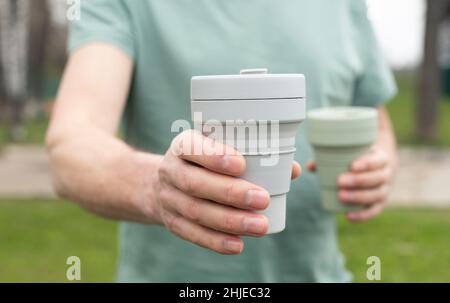 Mano maschio con due tazze eco riutilizzabili in mano, dando e offrendo tazza di caffè in silicone alla fotocamera. Foto Stock