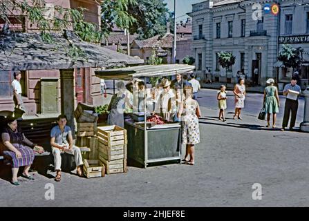 Una fotografia del 1960s di un commerciante di strada femminile che vende frutta e verdura da una stalla coperta sul manto stradale nel sole estivo di Mosca, Russia. Spesso i giardinieri di mercato e i proprietari di piccole aziende provenienti da fuori della città sarebbero venuti in città ogni settimana per vendere i loro prodotti coltivati in casa. Qui sembra che le prugne e le mele siano l'attrazione principale per la fila di acquirenti. Questa immagine proviene da una trasparenza amatoriale del colore Agfacolor 35mm – una fotografia d'epoca del 1960s. Foto Stock