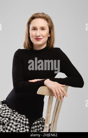 Donna sorridente in dolcevita seduta su sedia alta appoggiata alla schiena in studio Foto Stock