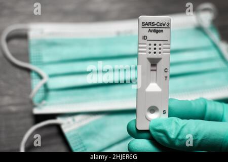 Mano con guanto usa e getta contiene il kit di test per coronavirus con maschere per il viso sullo sfondo. L'autotest dell'antigene rapido mostra un risultato negativo Foto Stock