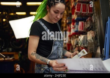 Coltivare giovane dipendente informale femminile con tatuaggio sul braccio e lungo verde dreadlocks leggere le informazioni nel giornale mentre in piedi al tavolo durante il lavoro in Foto Stock