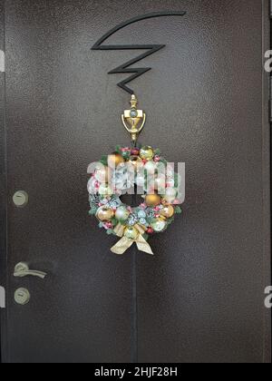 La lussuosa corona decorativa natalizia, fatta a mano con coni di pino e palle dorate, è appesa alla porta anteriore scura della casa. Foto Stock