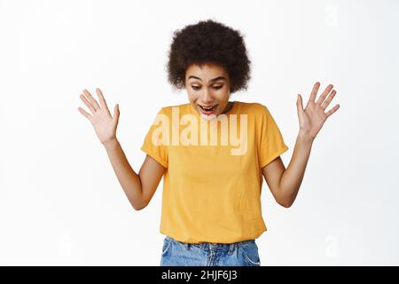 Ritratto di sorpresa donna africana alzando le mani in su, guardando giù in stupore e stupore, vedere qualcosa di emozionante sotto, urlare con entusiasta Foto Stock