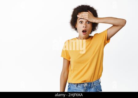 Scioccata ragazza nera schiaffa la fronte, guardando allarmato e preoccupato, caduta mascella e fissando insicuro alla macchina fotografica, indossando la t-shirt gialla, sfondo bianco Foto Stock