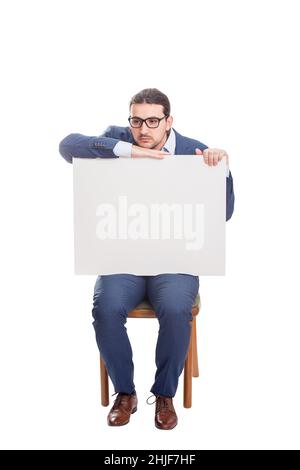 Uomo d'affari malinconico seduto su sedia con un grande banner vuoto con spazio per i messaggi. Persona di affari di lunghezza completa che osserva pensieroso da parte. Foto Stock