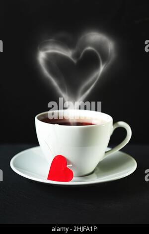 Tazza bianca di tè con fumo a forma di cuore. Sfondo nero, messa a fuoco selettiva, tonalità. Foto Stock