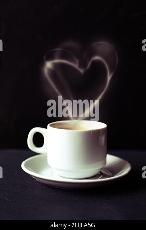 Tazza bianca di caffè espresso con fumo a forma di cuore. Sfondo nero, messa a fuoco selettiva, tonalità. Foto Stock
