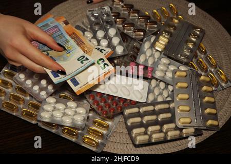 La mano femminile tiene le banconote in euro su molti blister di compresse, farmaci, pillole al tavolo Foto Stock