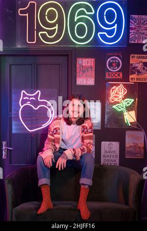 Kiev, Ucraina - 16 dicembre 2021: Uomo millenario in stile retrò che balla e salta sul divano. Nostalgia e illuminazione interna al neon. Foto Stock