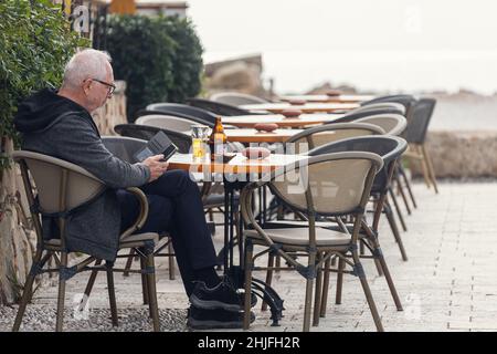 Side, Turchia -22 gennaio 2022: Uomo anziano che riposa in un caffè, leggendo un libro e bevendo birra in vacanza Foto Stock