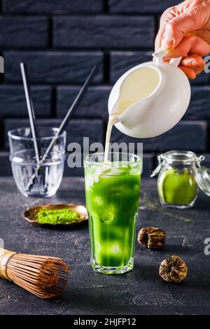 Barista versare il latte a mano in un bicchiere con tè Iced Green Matcha o latte match mescolato con cubetti di ghiaccio con una parete di mattoni sullo sfondo e in polvere m Foto Stock