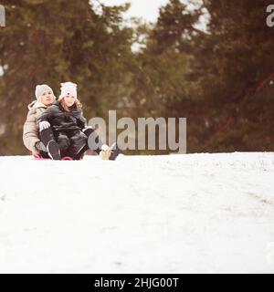 Madre felice con bambina seduta su slitta e slittino giù sulla neve dalla collina. Goditi una giornata invernale bianca al parco. Passare del tempo insieme Foto Stock