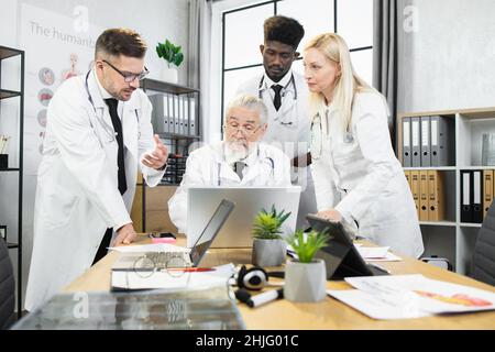 Gruppo di quattro medici specialisti multietnici che hanno una conferenza in ufficio. Medico capo anziano seduto al tavolo con un computer portatile mentre i colleghi sono in piedi intorno. Foto Stock