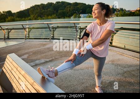 Donna atletica sicura e determinata, pareggiatore, runner in abbigliamento sportivo, cuffie, in piedi su un tapis roulant, mettendo le gambe su panca di legno Foto Stock
