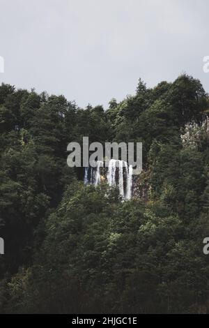 Un angolo basso di una cascata sulla montagna circondata da alberi contro un cielo nuvoloso Foto Stock