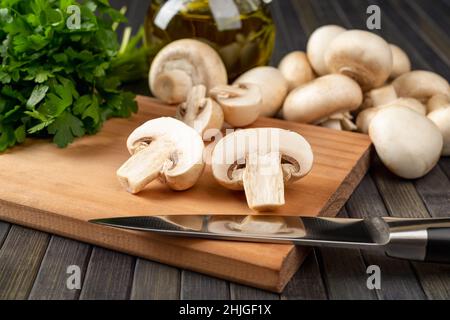 Champignon crudo tagliato a metà e coltello da cucina su un tagliere, funghi a bottoni interi e un mazzo di prezzemolo su sfondo scuro. Cibo vegetariano Foto Stock