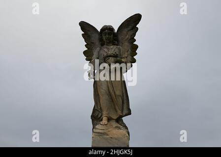 Scultura in pietra antica e usurata di un angelo in lutto che cade fiori, con un cielo grigio e nuvoloso sullo sfondo Foto Stock