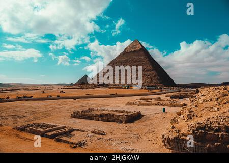 Grandi piramidi di Giza in una soleggiata giornata invernale, diversi autobus turistici in lontananza. Cielo nuvoloso blu brillante, sabbia arancione vibrante che copre le rovine Foto Stock