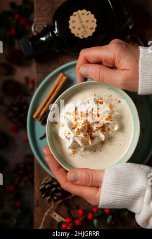 Un paio di mani intorno a una tazza di latte di pan di zenzero con panna montata e biscotti sbriciolati. Foto Stock