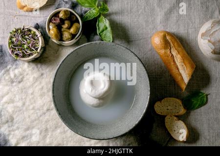 Burrata tradizionale italiana annodata formaggio in ceramica grigia ciotola sul tavolo. Pane, olive, insalata verde intorno. Ingredienti per un sano din mediterraneo Foto Stock