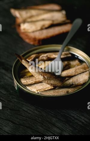 Lattina aperta con spratti baltici affumicati in olio, uno sulla forchetta, con fetta di pane di segale dietro su tavola di legno nero. Primo piano Foto Stock