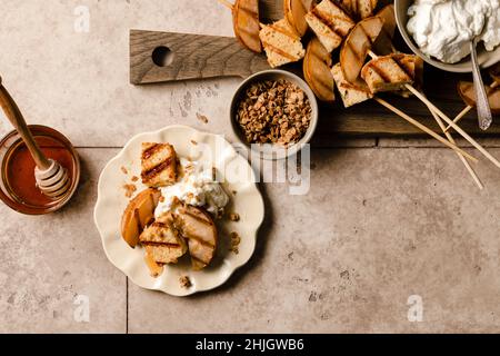 Torta di sterlina alla griglia e pere su un piatto con panna montata, miele, e una spruzzata di granola accanto a un bordo di servizio di spiedini cake di sterlina alla griglia e Foto Stock