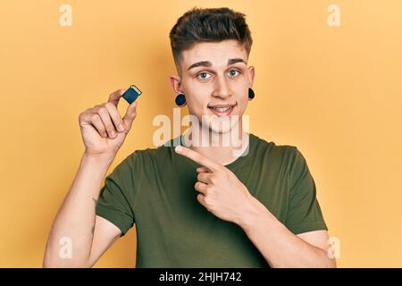 Giovane ragazzo caucasico con dilatazione delle orecchie che tiene la scheda sdxc sorridendo felice puntando con mano e dito Foto Stock