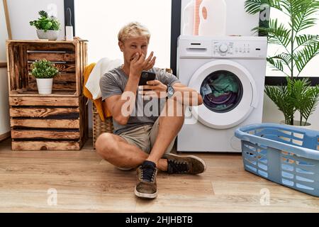 Giovane bionda che fa lavanderia usando lo smartphone che odora qualcosa di stinky e disgustoso, odore intollerabile, che tiene il respiro con le dita sul naso. Male s Foto Stock