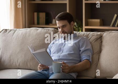 Sorridente giovane uomo rilassato coinvolto nella lettura libro di carta. Foto Stock