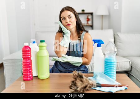 Giovane donna bruna che indossa grembiule più pulito e guanti pulizia a casa pensando di guardare stanco e annoiato con problemi di depressione con le braccia incrociate. Foto Stock