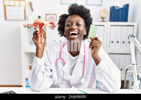 Medico africano donna che tiene anatomico organo genitale femminile e pillole di controllo delle nascite che celebra pazzo e stupito per il successo con occhi aperti urlando Foto Stock