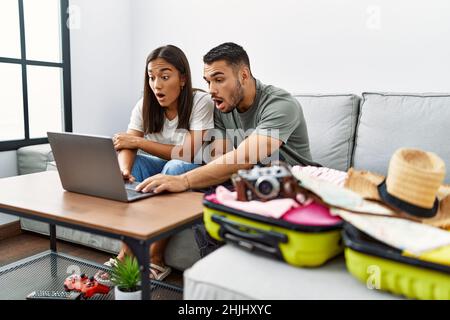 Giovane coppia interrazziale che impacchettano abiti estivi in valigia guardando il laptop spaventato e stupito con bocca aperta per sorpresa, faccia incredulità Foto Stock