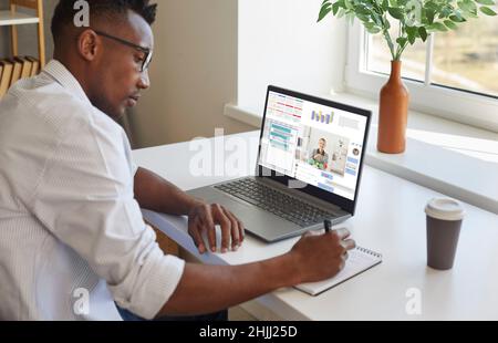 Uomo nero che lavora sul laptop, comunica tramite chat e videochiamata e prende appunti Foto Stock