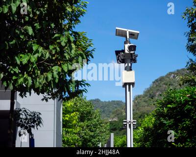 Moderna telecamera CCTV bianca sistema di sicurezza elettronico con pannello solare e luce stradale sul palo al passaggio pedonale nel resort vicino a alberi verdi Foto Stock