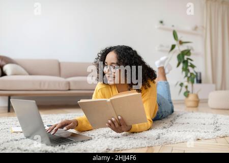 Giovane donna nera che legge il libro vicino al portatile, preparandosi per l'esame o lavorando al progetto, adagiata sul pavimento a casa Foto Stock
