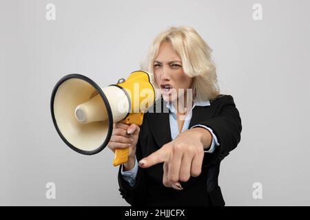 Agitato donna d'affari in abbigliamento formalwear gridando nel megafono e puntando il dito, posando su sfondo grigio Foto Stock