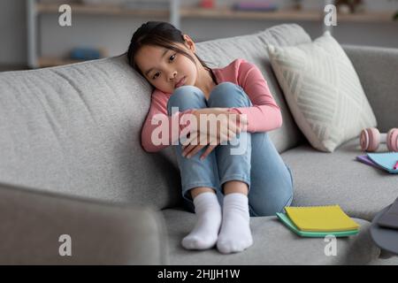 Scolaro asiatico sconvolto seduto sul divano, studiando a casa Foto Stock