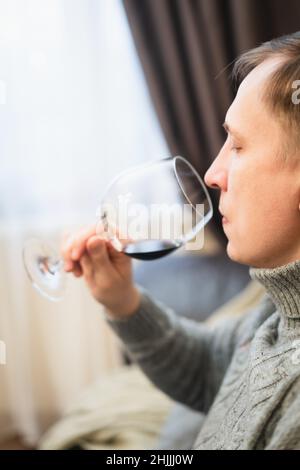 Rilassato sereno ricco solitario di mezza età single uomo bere vino rosso rilassarsi sul divano a casa, godendo di riposo seduto sul divano che tiene degustazione di vetro Foto Stock